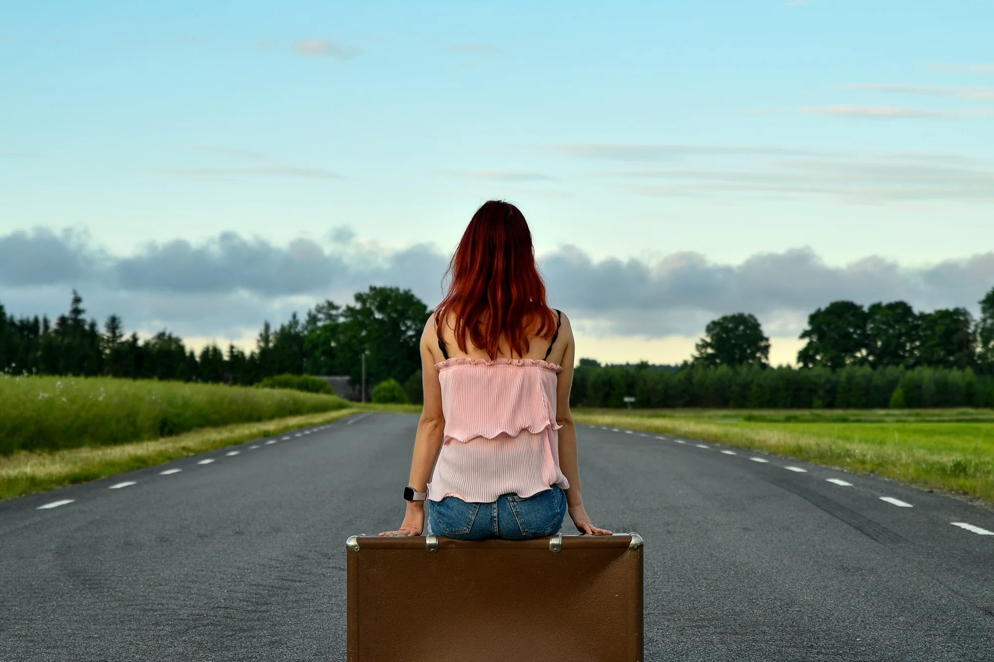 A woman sits alone on a suitcase in the middle of an empty road, her back to the camera, facing the horizon