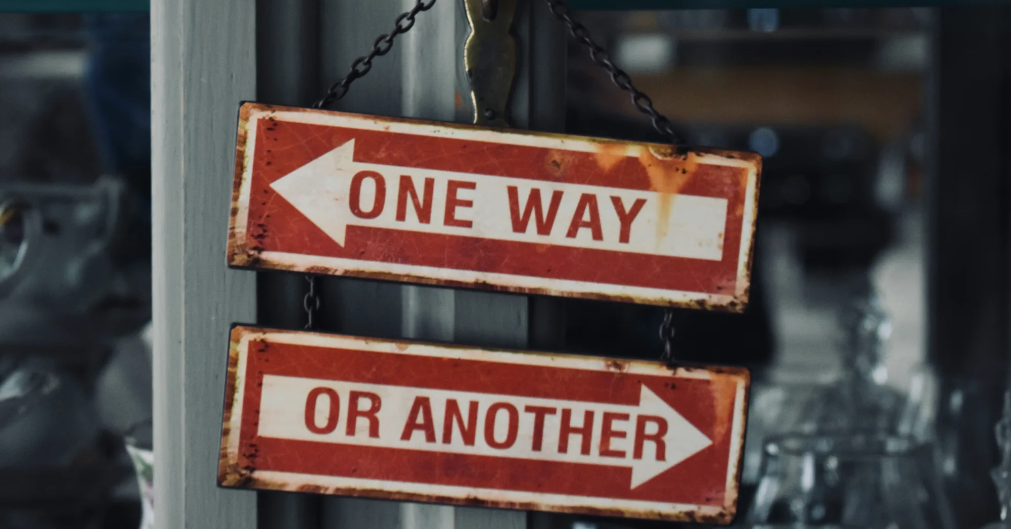 Two weathered red enamel signs hanging on a chain, one arrow pointing left reading ONE WAY, the other pointing right reading OR ANOTHER